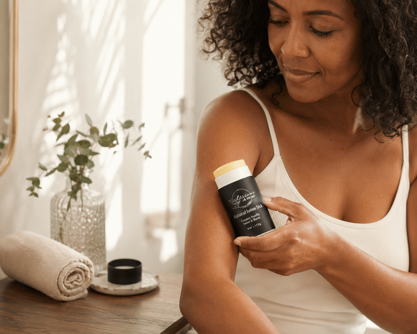 A woman with curly dark hair applies a creamy vanilla lotion stick to her upper arm. The product container is black with a light yellow cap, held by her hand near a wooden table with a rolled towel and a vase of greenery in the background.