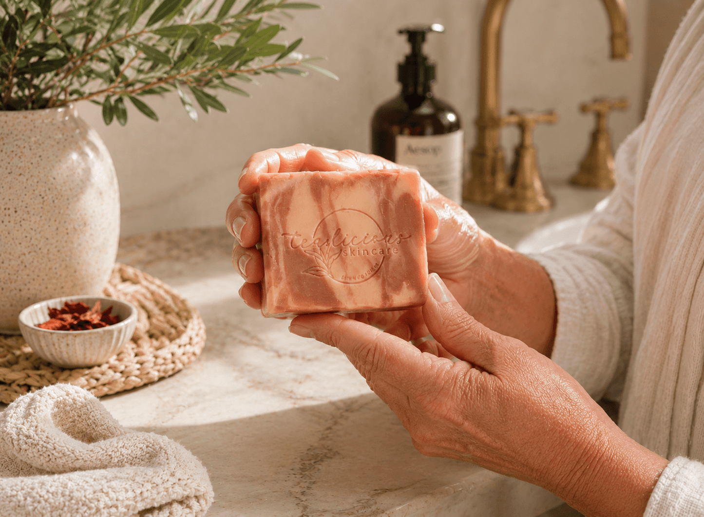 Woman with naturally textured hands holding a handcrafted Moroccan Cashmere cold process soap bar in a sunlit luxury bathroom, featuring warm marble tones, brass fixtures, botanical accents, and a soft spa-inspired setting.