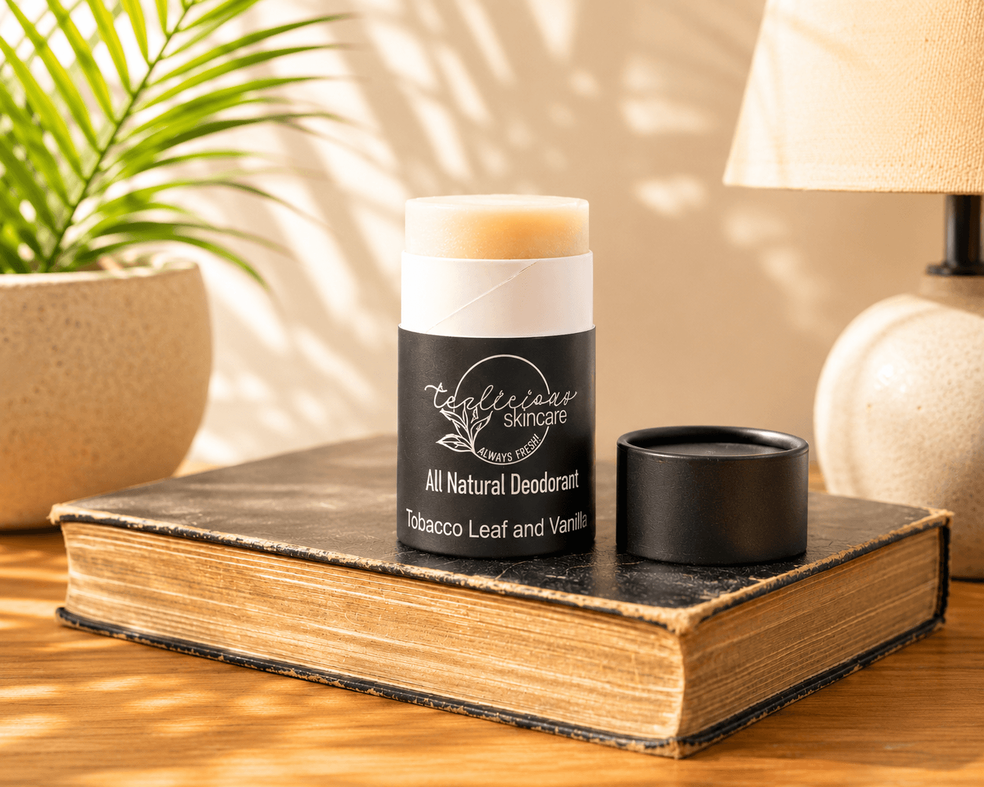 A deodorant stick and its cap sit on an old book on a wooden table, styled with a potted plant and a lamp in the background.