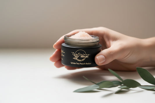 A person’s hand holding an open jar of Tea-licious Skincare White Tea Peach Bellini lip scrub, the granular texture of the botanical-infused formula. The shot follows a Professional Homestyle aesthetic with soft lighting and a sprig of green leaves on a white surface.