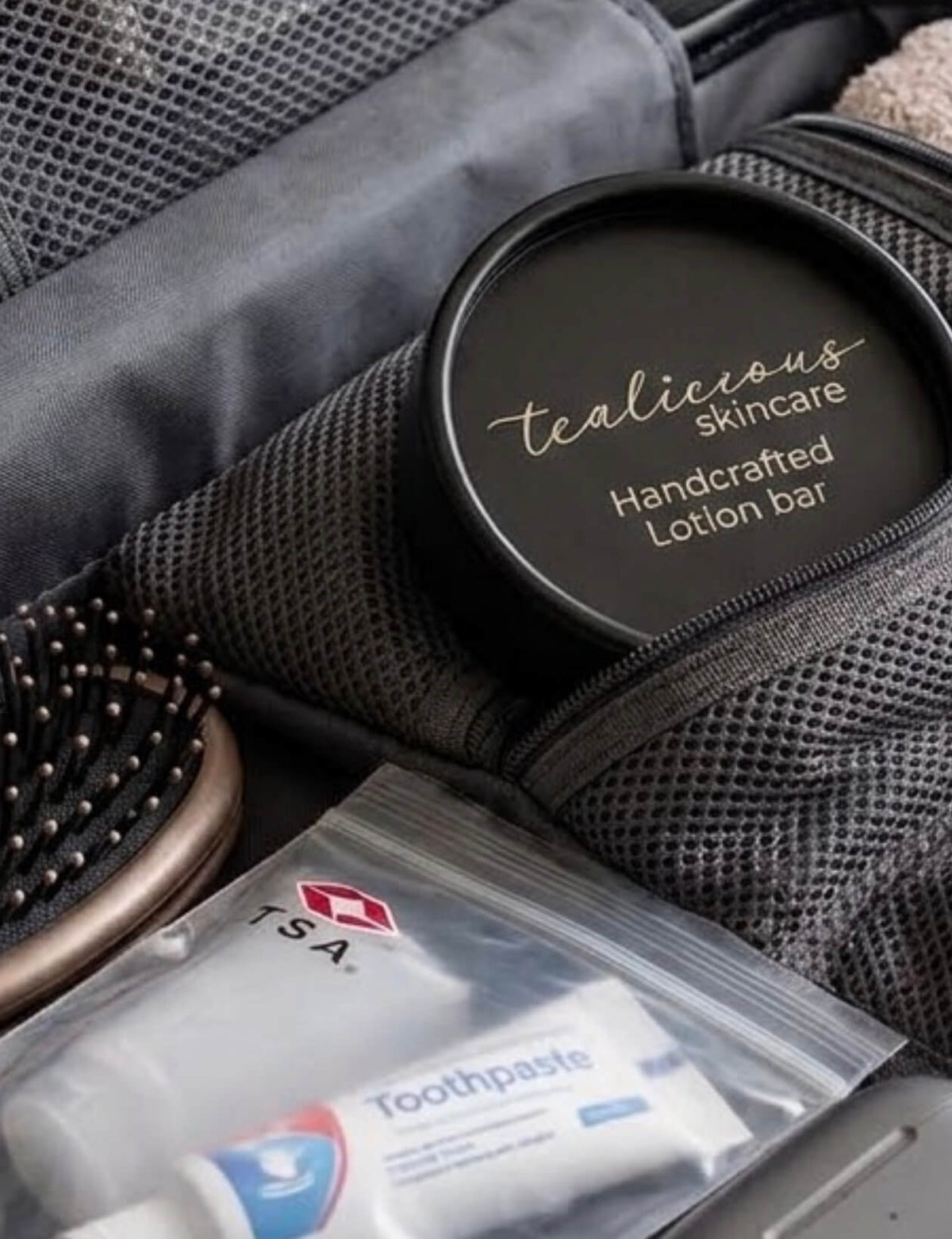 Tealicious Skincare handcrafted solid lotion bar in a black screw-top tin with metallic silver lettering, packed securely inside an open carry-on suitcase alongside travel essentials.