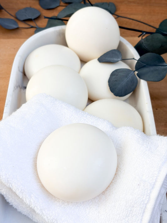 A white bowl lined with a white towel, filled with smooth, round, creamy white soaps—perfectly formed, unscented, and minimalist. Surrounding a few scattered eucalyptus leaves.