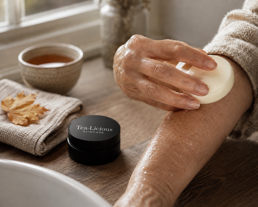 Close-up of a person gliding a round, white solid lotion bar across their forearm. In the background on a wooden table, a small black Tea-Licious Skincare container sits beside a towel, an autumn leaf, and a cup of brewed tea by a window