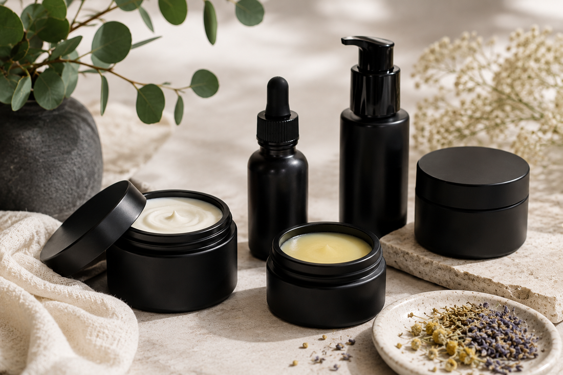Matte black skincare jars and bottles arranged with botanical ingredients on a neutral stone surface in soft natural light, representing premium small-batch skincare.