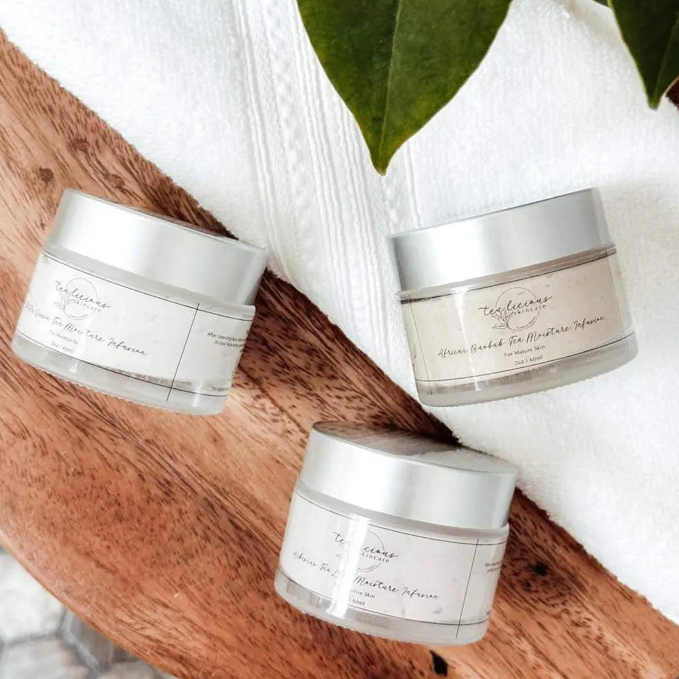 Three elegant jars of Tea-licious Skincare Moisture Infusions—including Hibiscus Tea and African Baobab Tea—beautifully arranged on a soft white towel with a fresh green leaf and warm wooden accents