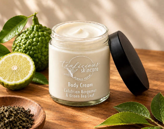 Open clear glass jar of Tea-licious Skincare Calabrian Bergamot & Green Tea Leaf Body Cream filled with smooth white cream, set on a warm wooden surface beside fresh bergamot fruit, whole green tea leaves in a small wooden dish, and a black lid.