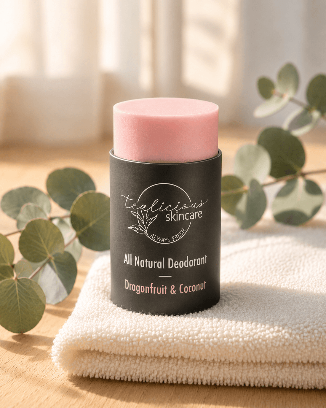 Tea-licious Skincare Dragonfruit & Coconut natural deodorant stick in a black container, sitting on a white towel with a sprig of eucalyptus in the background, lit by warm, natural morning sunlight