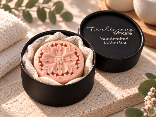 A round pink lotion bar with an embossed floral pattern sits in a black round tin lined with beige fabric. The black lid, printed with 'Tealicious Skincare Handcrafted Lotion bar,' rests beside the tin on a textured beige surface with soft natural shadows and decorative greenery.