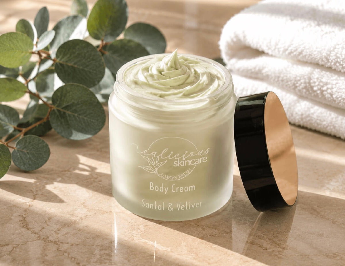 Tea-licious Skincare jar of Santal and vetiver body cream with its lid off, placed on a marble surface with eucalyptus leaves and a white towel in the background. The jar is frosted with pale green cream visible inside, and the lid has a black exterior and gold interior.