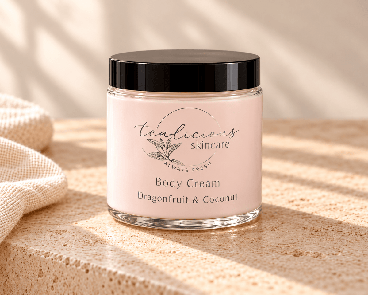 A jar of tealicious skincare Tea-licious dragonfruit coconut body cream with a black lid and pale pink cream inside, set on a textured beige stone surface with soft natural shadows.