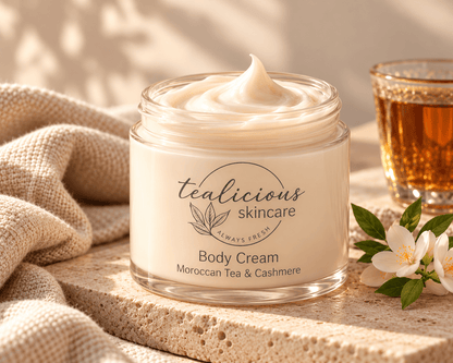 A jar of Tealicious Skincare Moroccan Tea & Cashmere body cream sits on a stone surface with a beige linen cloth, a glass of tea, and white flowers around it.