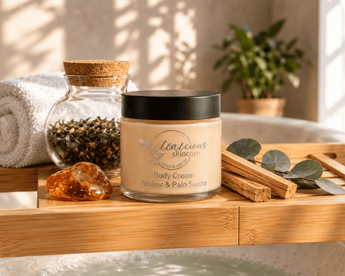 Tea-licious Amber & Palo Santo Body Cream on a bamboo bath tray beside loose tea, a white towel, amber resin, Palo Santo wood and eucalyptus.