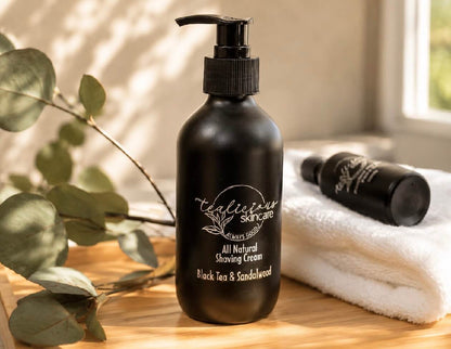 Tea-infused Black Tea & Sandalwood shaving cream in a black pump bottle displayed on a wooden countertop with a white towel and eucalyptus leaves.