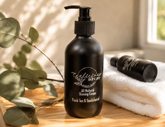 Tea-infused Black Tea & Sandalwood shaving cream in a black pump bottle displayed on a wooden countertop with a white towel and eucalyptus leaves.
