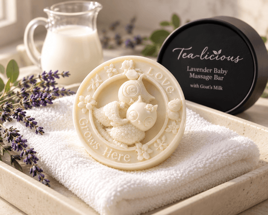 Tea-licious Lavender Baby Massage Bar with Goat’s Milk on a white towel beside its black container, lavender sprigs and a milk pitcher.