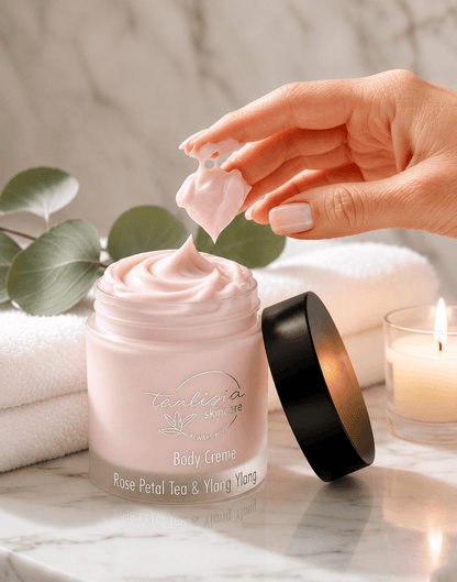 Hand applying tea-licious skincare rose petal tea and lay ylang cream from a jar labeled 'Tealicious Skincare' on a marble surface with a candle and eucalyptus leaves in the background.