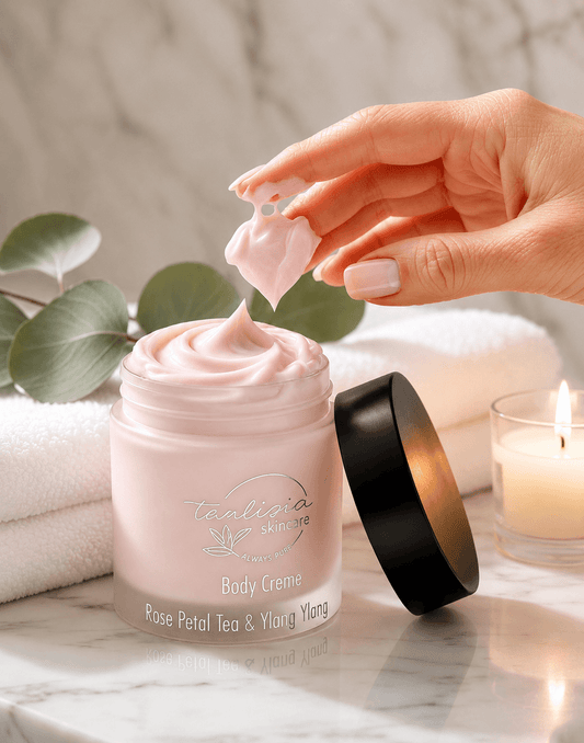 Hand applying tea-licious skincare rose petal tea and lay ylang cream from a jar labeled 'Tealicious Skincare' on a marble surface with a candle and eucalyptus leaves in the background.