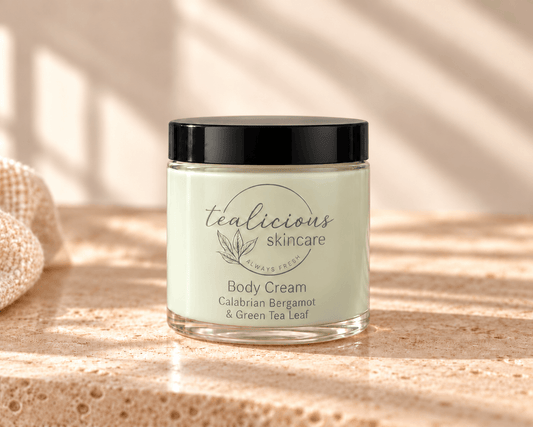 A jar of tealicious skincare Calabrian Bergamot & Green Tangerine Signature Hybrid Body Cream sits on a textured stone surface with warm sunlight and shadows. The jar has a black lid and a pale green label with the brand name 'tealicious skincare' and product details for 'Body Cream' in Calabrian Bergamot & Green Tea Leaf.