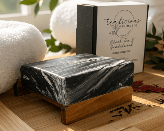 Tea-licious Black Tea & Sandalwood tea-infused men’s soap bar with black and white marbled swirls, displayed on a wooden soap dish beside its product box in a bright spa-inspired setting.