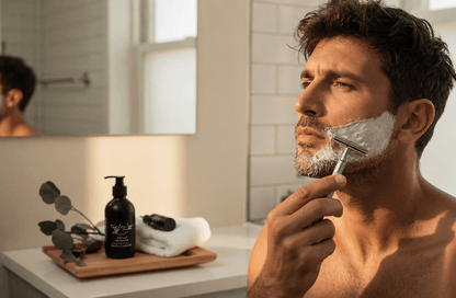 Man applying Black Tea & Sandalwood tea-infused shaving cream with a safety razor in a bright bathroom, with Tea-licious Skincare shaving cream displayed on the vanity.
