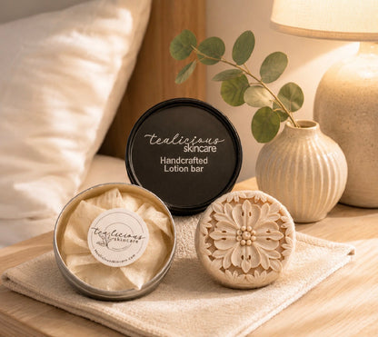 Tealicious Skincare handcrafted lotion bar shown in a styled bedroom setting, with the round black lid, an open tin showing the beige lotion bar, and a carved wooden bar on a beige towel.