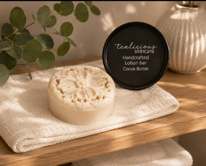 Cocoa Butter & Cashmere tea-infused lotion bar with handcrafted floral design beside its tin on a folded towel, providing concentrated waterless moisture for dry skin.