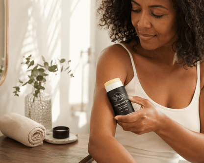 A woman with curly dark hair applies a creamy vanilla lotion stick to her upper arm. The product container is black with a light yellow cap, held by her hand near a wooden table with a rolled towel and a vase of greenery in the background.