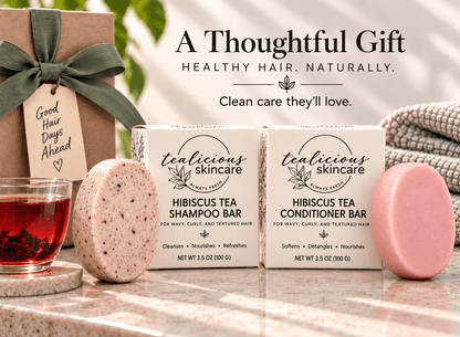 Tealicious Skincare Hibiscus Tea Shampoo Bar and Conditioner Bar gift set displayed with their packaging boxes, a wrapped gift box, a glass of red tea, and a pink shampoo bar on a marble surface.