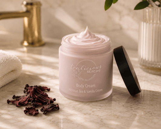 A jar of 'tenacious skincare' body cream with its lid off, showing the whipped cream inside. The jar is placed on a marble surface next to dried hibiscus flowers, with a gold faucet and a towel blurred in the background.