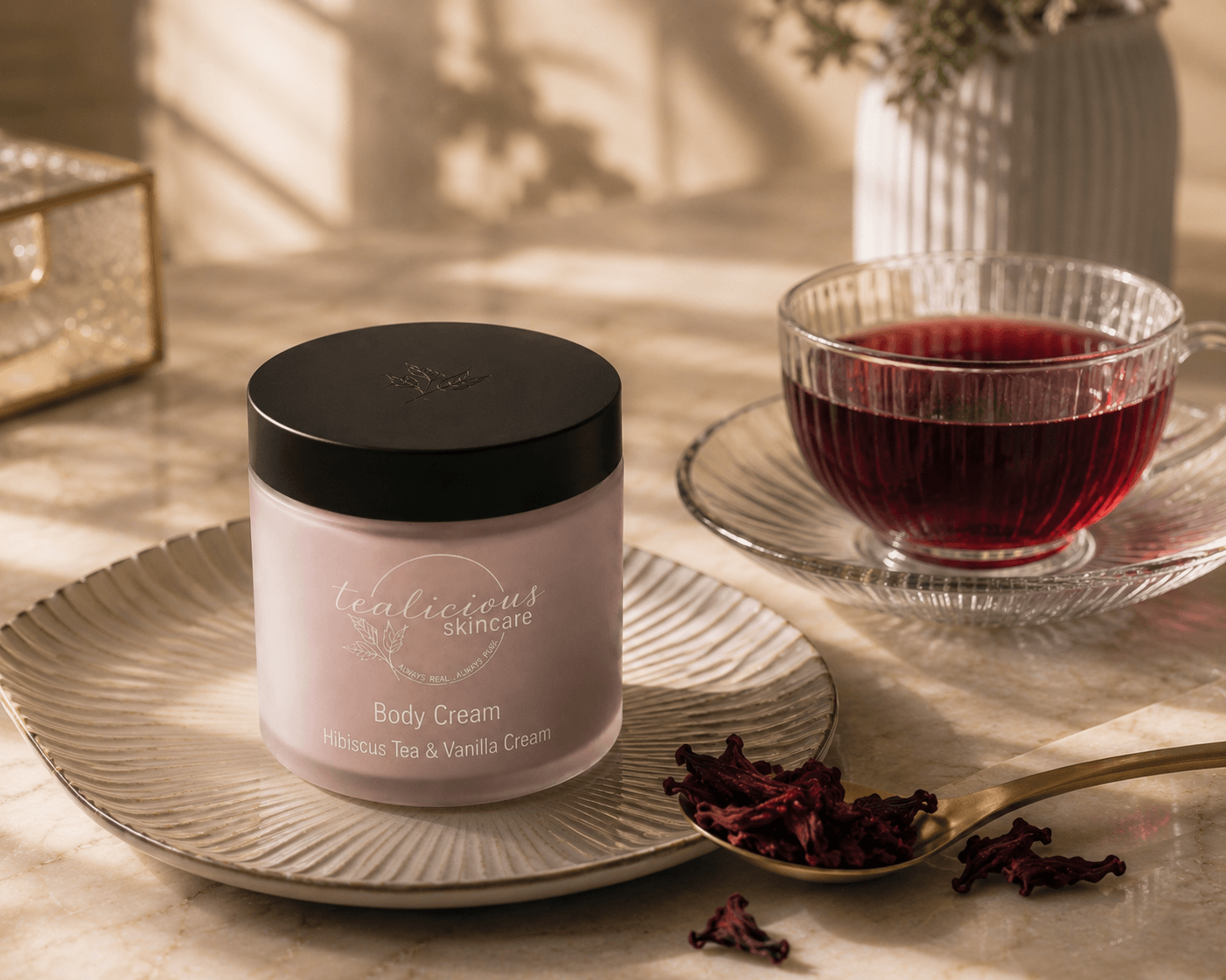 A jar of luxurious skincare body cream sits on a patterned plate, styled with a glass of red tea and dried hibiscus flowers on a marble surface.