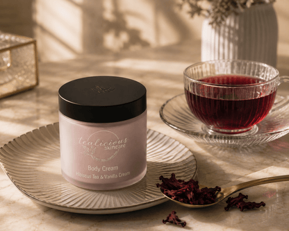 A jar of luxurious skincare body cream sits on a patterned plate, styled with a glass of red tea and dried hibiscus flowers on a marble surface.