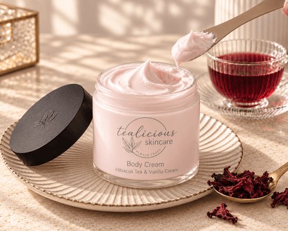 Tealicious Skincare jar of luxurious hibiscus tea and vanilla body cream showing texture on a patterned plate, styled with a glass of red tea and dried hibiscus flowers on a marble surface.