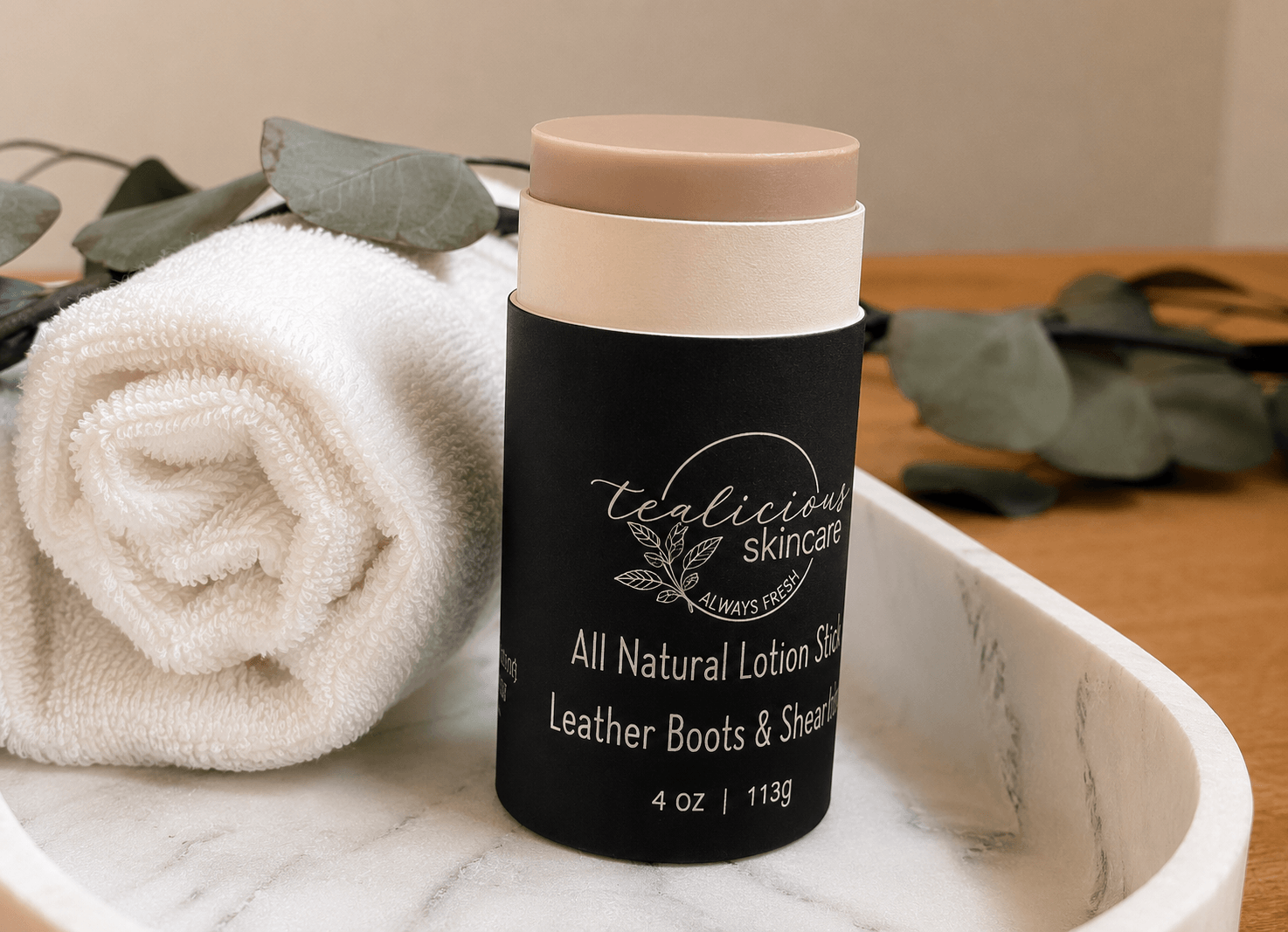 Tea-Licious Skincare Leather Boots & Shearling natural lotion stick on a marble tray with a white towel and eucalyptus in a warm spa-inspired bathroom setting.