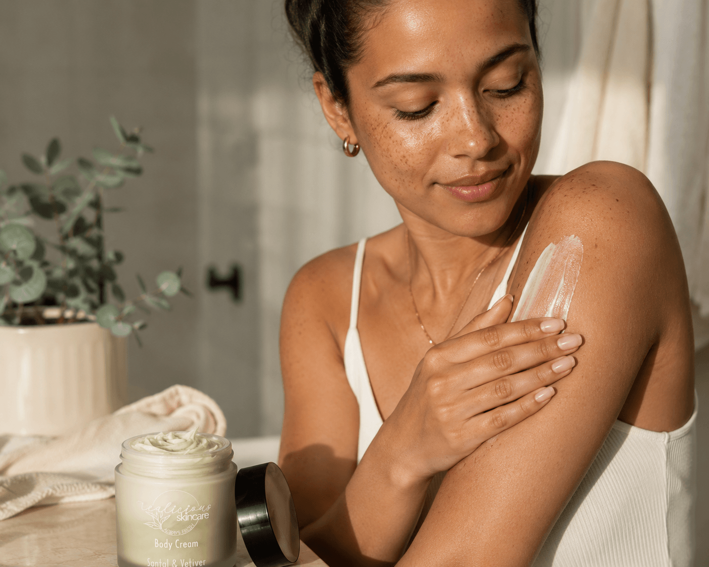 A woman applies a pale green Santal and vetiver body cream from a jar labeled onto her upper arm in a warm, sunlit indoor setting.