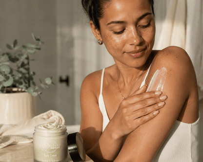 A woman applies a pale green Santal and vetiver body cream from a jar labeled onto her upper arm in a warm, sunlit indoor setting.