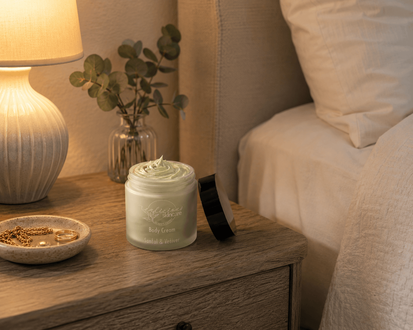 A jar of pale green body cream with its lid off, placed on a wooden bedside table next to a lit lamp, a small vase with eucalyptus, and a bed with white linens.