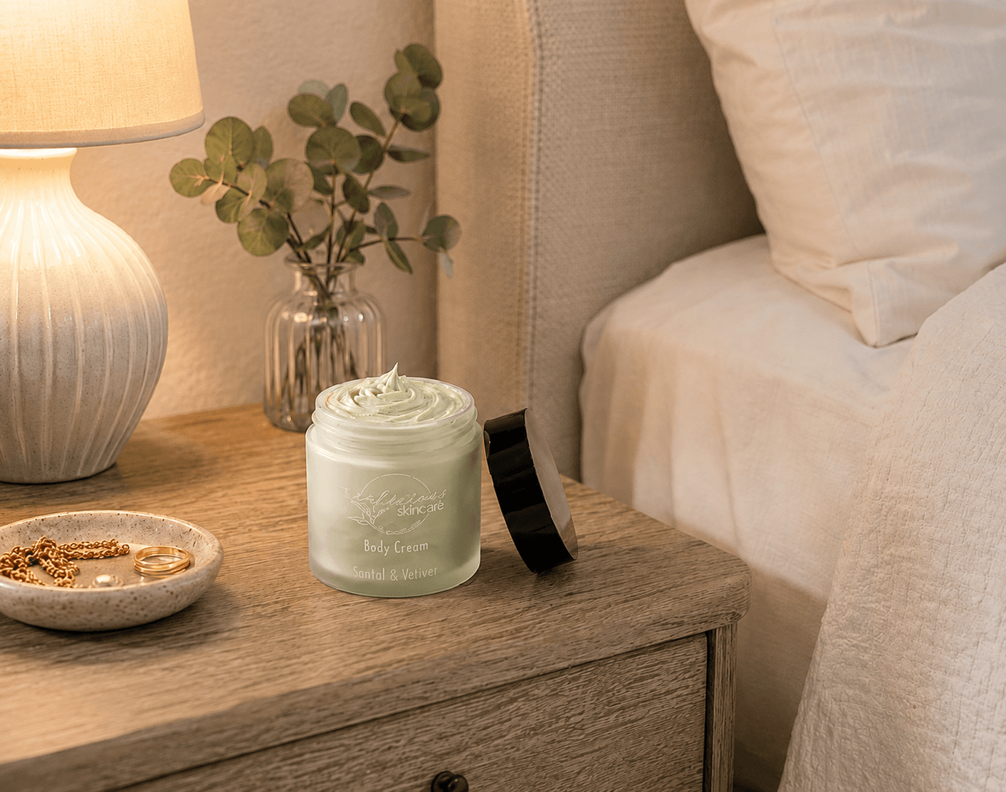 Tea-infused Santal & Vetiver body cream in an open glass jar on a wooden bedside table beside a lamp, eucalyptus, and white bedding, showcasing rich, whipped hydration.