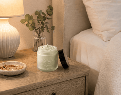 Tea-infused Santal & Vetiver body cream in an open glass jar on a wooden bedside table beside a lamp, eucalyptus, and white bedding, showcasing rich, whipped hydration.