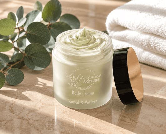 A jar of Santal and vetiver body cream with its lid off, placed on a marble surface with eucalyptus leaves and a white towel in the background. The jar is frosted with pale green cream visible inside, and the lid has a black exterior and gold interior.