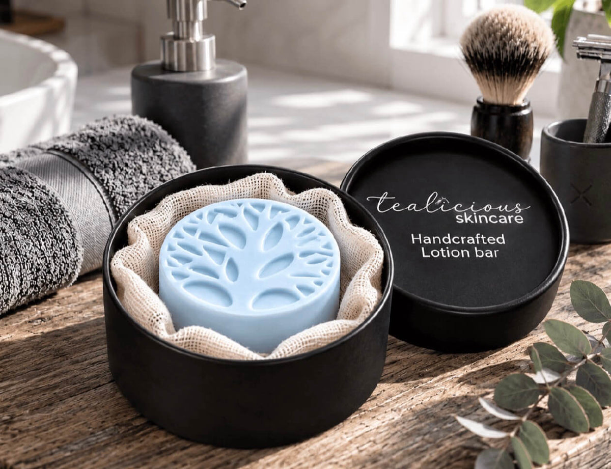A tealicious lotion bar container with its lid beside it, showing a light blue lotion bar with a tree design embossed on top. The container is set on a rustic wooden surface with spa-style bathroom props including a towel, soap dispenser, shaving brush, and razor.