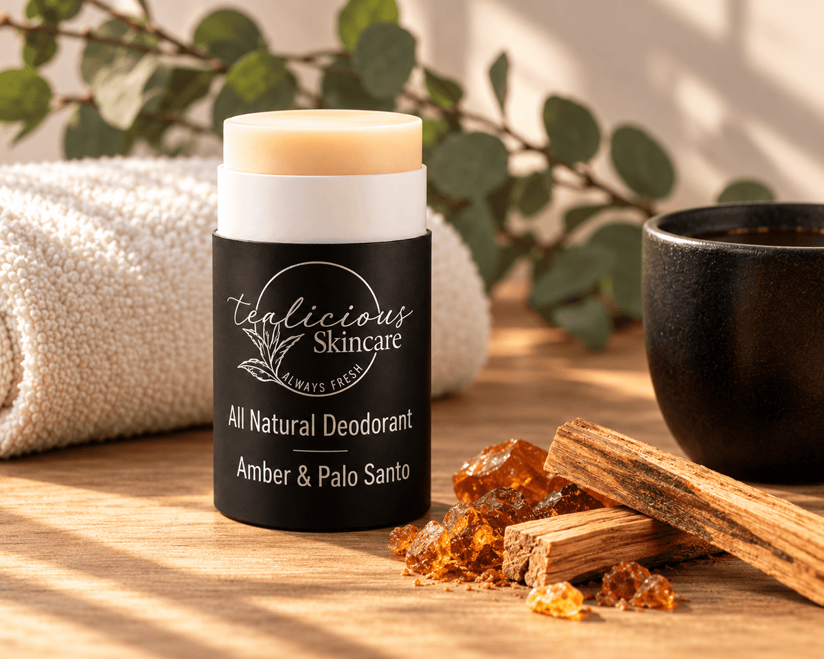 Tea-licious Amber & Palo Santo deodorant on a wooden surface beside amber-colored resin and wood sticks, with a cream towel and greenery in warm sunlight.