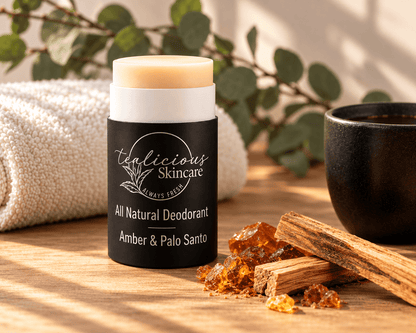 Tea-licious Amber & Palo Santo deodorant on a wooden surface beside amber-colored resin and wood sticks, with a cream towel and greenery in warm sunlight.