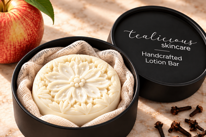 Tea-Licious handcrafted floral solid lotion bar in a matte black tin beside a red apple and whole cloves on a warm stone surface.