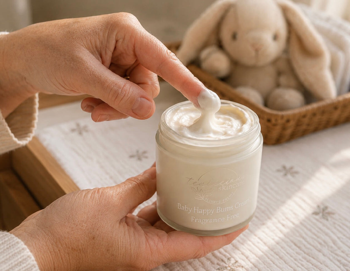 A hand scoops fragrance-free Baby Happy Bums Cream from its jar, with a plush bunny in the softly lit background.