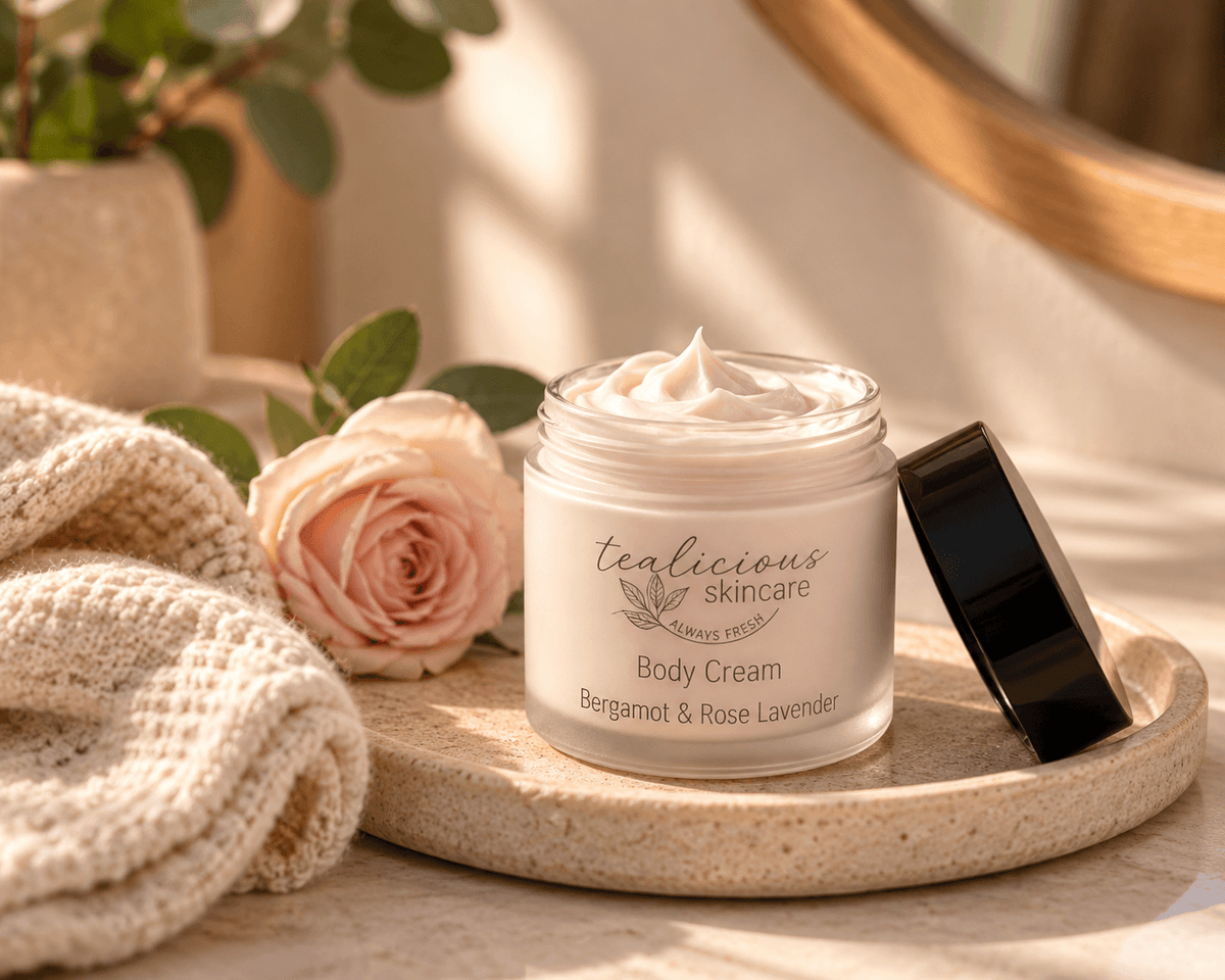 Tea-Licious Bergamot & Rose Lavender Body Cream in an open glass jar on a stone tray beside a soft pink rose and cream towel.