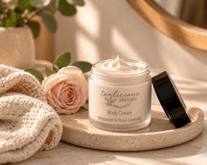 Tea-Licious Bergamot & Rose Lavender Body Cream in an open glass jar on a stone tray beside a soft pink rose and cream towel.