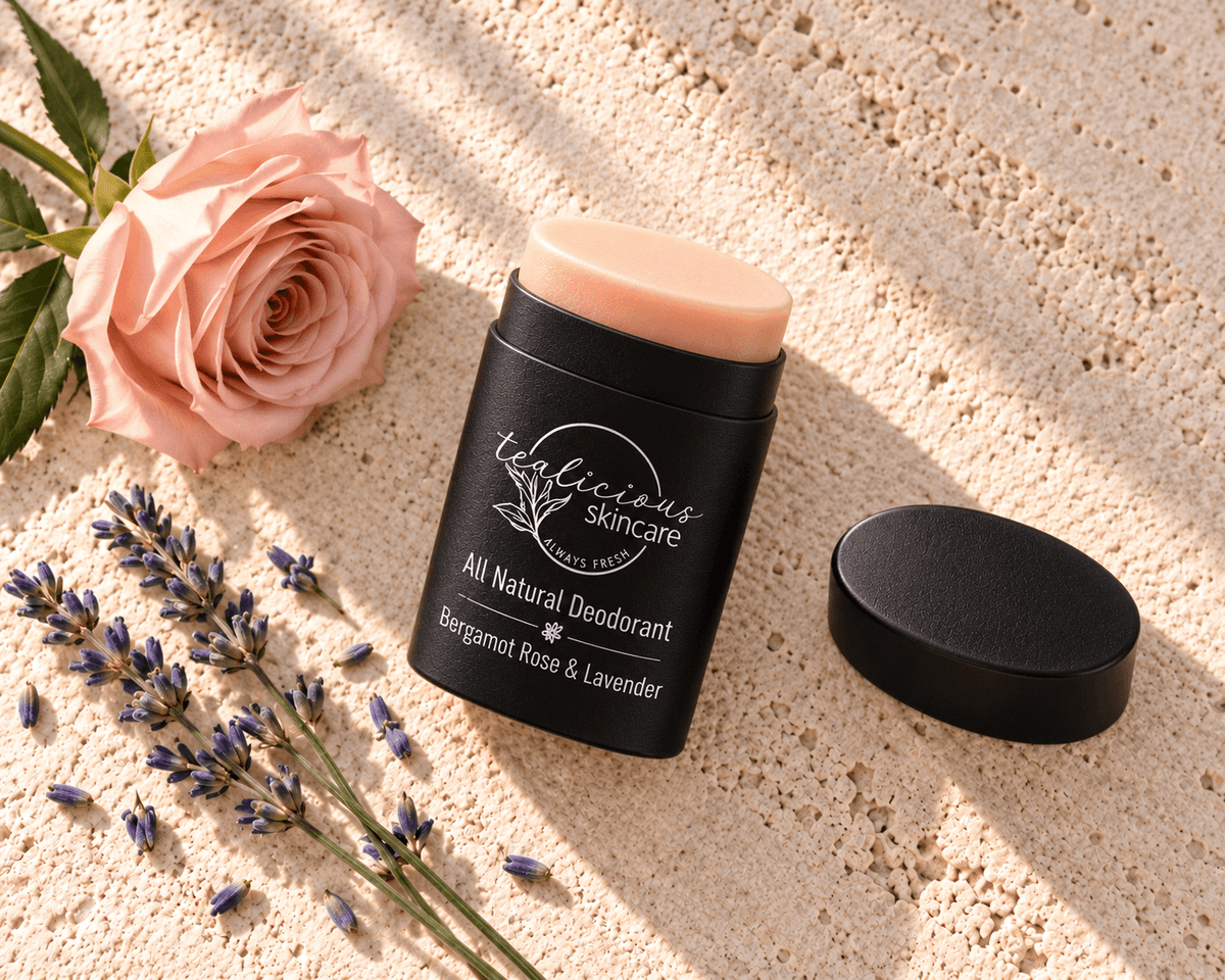 Tea-licious Bergamot Rose & Lavender deodorant in a black oval container with silver lettering, beside its lid, a pink rose and lavender on cream stone.
