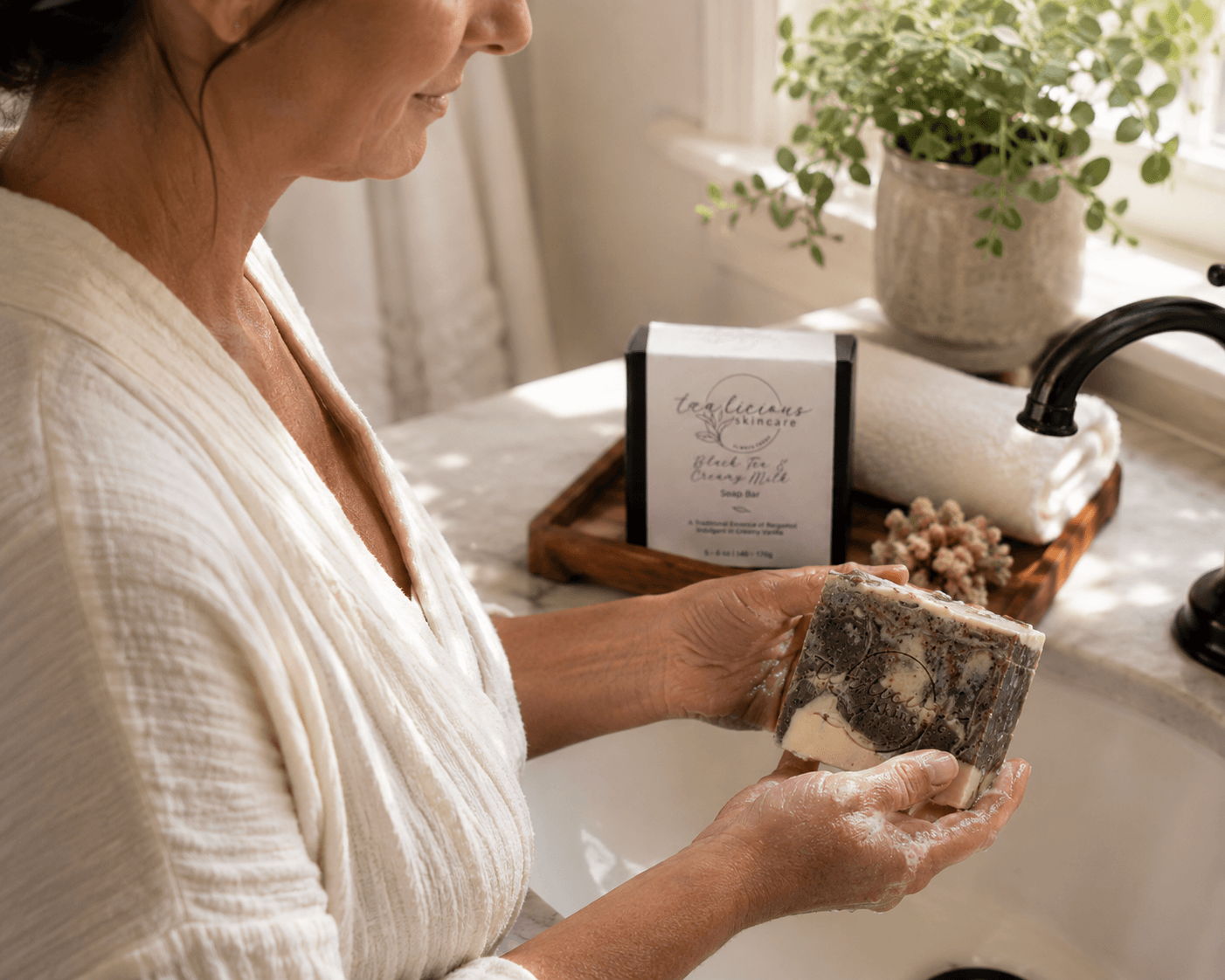 Woman holding Tea-Licious Black Tea & Creamy Milk tea-infused soap bar over a bathroom sink with soft natural morning light and real skin texture.