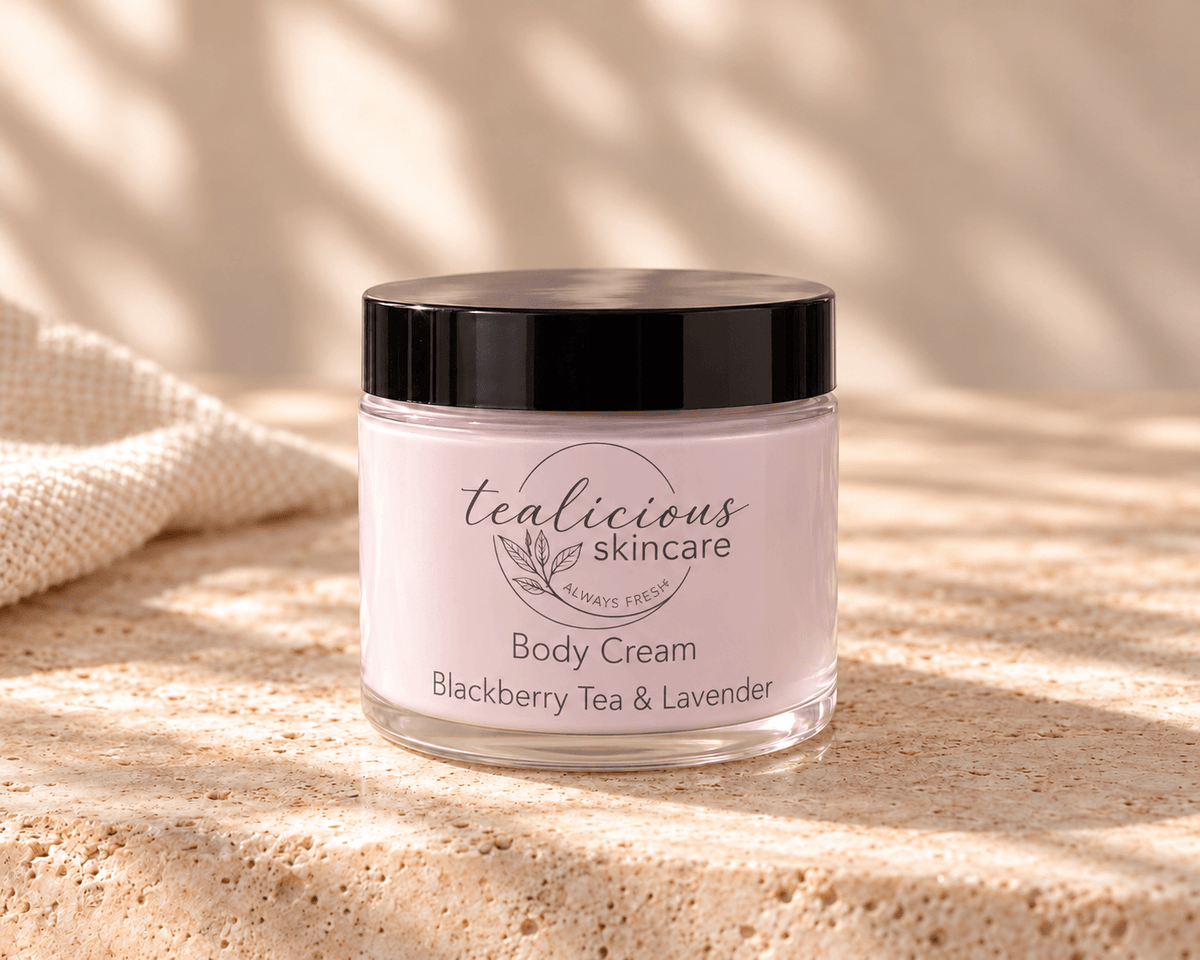 Tea-licious Blackberry Tea & Lavender Body Cream in a glass jar with a black lid, on neutral stone beside cream linen in soft natural light.