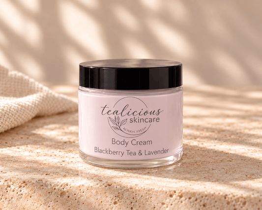 Tea-licious Blackberry Tea & Lavender Body Cream in a glass jar with a black lid, on neutral stone beside cream linen in soft natural light.
